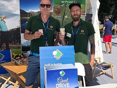 Zwei Männer in grünen Polohemden stehen lächelnd vor einem Stand mit der Aufschrift "Golf in Austria" und halten Getränkebecher. Hintergrund mit Bergmotiv.