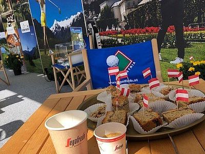 Ein Tisch mit Kuchenstücken in Papierförmchen, dekoriert mit kleinen österreichischen Fahnen. Im Hintergrund sind Berge und Blumen zu sehen.