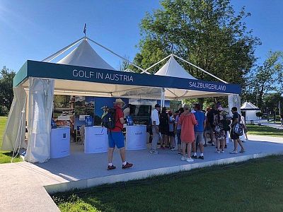 Outdoor booth with 'Golf in Austria' and 'Salzburgerland' banners, surrounded by visitors, sunny sky, green trees, and promotional materials visible.