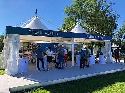 A promotional tent with 'Golf in Austria' signage. People interact with staff under a clear blue sky. Trees and green grass are visible in the background.