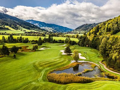 A lush green golf course with rolling hills, sand bunkers, and a pond. Mountains and a partly cloudy sky in the background create a picturesque view.