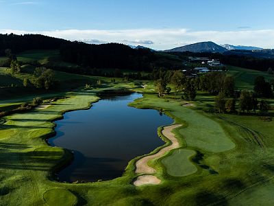 Aerial view of a golf course with a lake, lush green fairways, and surrounding hills under a clear blue sky.
