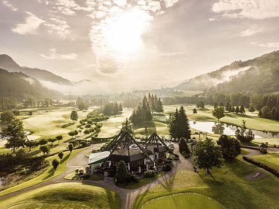 Aerial view of a scenic golf course with lush greenery, a clubhouse in the center, and rolling hills under a partly cloudy sky, creating a tranquil atmosphere.