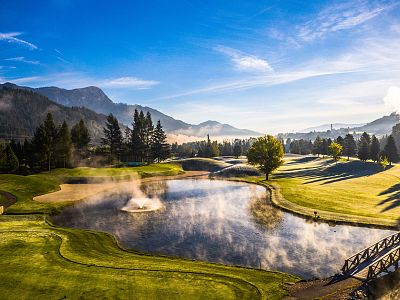 Scenic view of a golf course at sunrise, with mist over a pond, green fairways, and mountains in the background, under a clear blue sky.