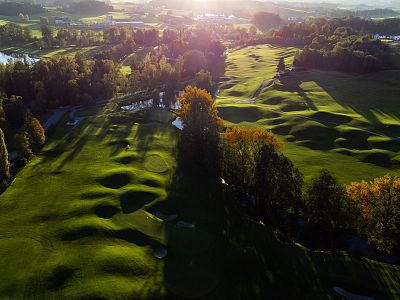 Aerial view of a golf course at sunset with lush greenery, shadowy hills, and sunlit trees creating a picturesque landscape in soft light.