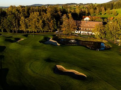 Aerial view of a golf course with lush fairways, a sand trap, a reflective pond, and a clubhouse surrounded by autumn trees under a setting sun.