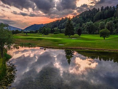 A serene landscape shows a golf course with lush greens, hills, and trees, under a sunset sky reflected in a still pond, creating a peaceful and scenic view.