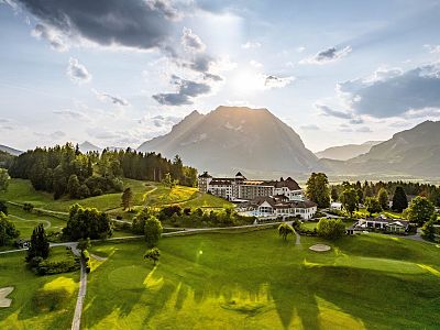A serene landscape with green hills, a large building, and mountains at sunset, under a partly cloudy sky.