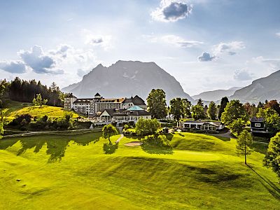 A grand hotel nestled in lush greenery, set against majestic mountains under a partly cloudy sky, offering a tranquil and scenic view.