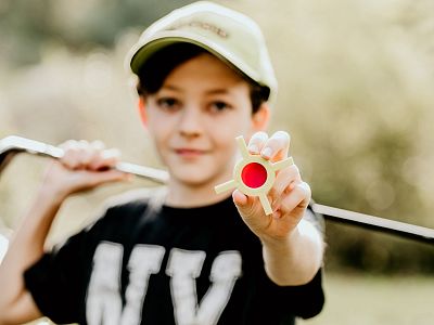 Young person wearing a cap holds a golf club over shoulder and shows a small red ball in their hand, suggesting a sporty outdoor scene.
