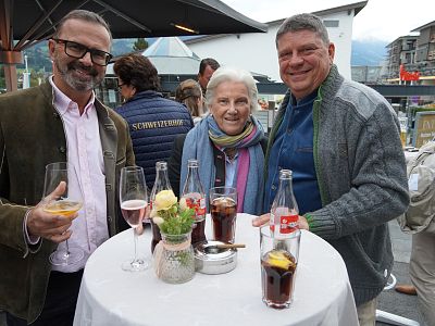 Three people stand around a small table outdoors, holding drinks and smiling. The table has glasses, a rose in a vase, and a small ashtray, set against a busy backdrop.