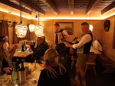 A group of people enjoy a meal in a cozy, wood-paneled room with traditional lights, while two musicians in Bavarian outfits play instruments and entertain them.