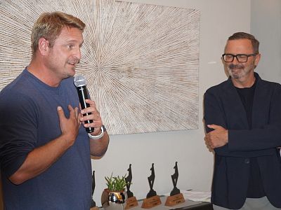 Two men at an event; one speaks into a microphone, the other listens. Award trophies and a wall art piece are in the background.