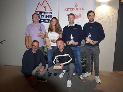 Six people pose with trophies in front of a backdrop featuring logos. Most are standing, holding awards, and smiling at the camera.
