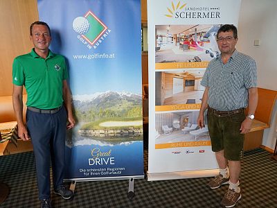 Two men stand beside banners promoting a golf event, with images of golf courses and accommodations, hinting at a scenic event setting.