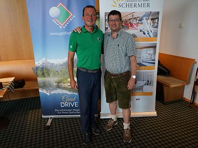 Two men pose in front of event banners, one in a green polo, the other in traditional attire, with a cozy indoor setting and promotional stands visible.