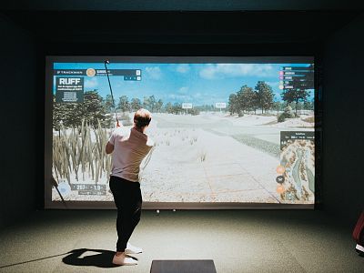 Person beim Indoor-Golfen in einem Simulatorraum, schwingt Golfschläger vor großer Leinwand, die eine virtuelle Golfanlage zeigt. Gedämpfte Beleuchtung.