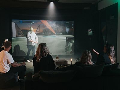 A group watching a video game projected on the wall in a dimly lit room with couches. Futuristic landscape is visible on the screen.