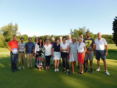 Eine Gruppe von Menschen steht auf einem Golfplatz. Sie lächeln in die Kamera, umgeben von grünen Bäumen und unter blauem Himmel. Es ist ein sonniger Tag.