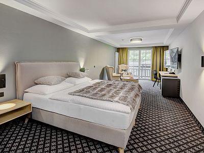 A hotel room with a large bed, dark carpet, armchair, desk, TV, and sliding glass doors to a balcony. The decor is contemporary and inviting.
