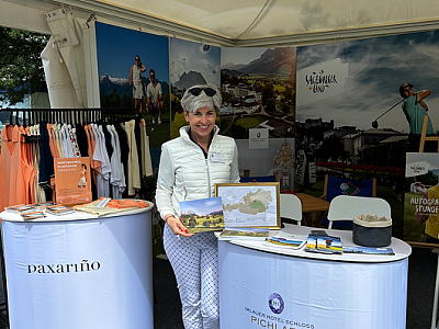 Person at a promotional booth with maps and brochures, showcasing travel information. Background includes landscape photos.