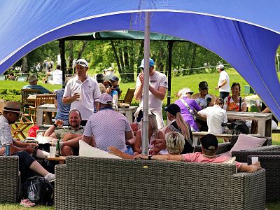A group of people sitting on sofas under a blue canopy, socializing in a sunny park with trees in the background.