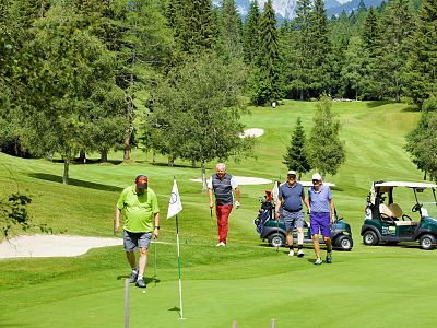 A group of golfers on a green course with trees, golf carts, and hills in the background on a sunny day.