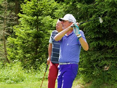 Two golfers in bright attire on a lush green course, one swinging the club while another observes near tall trees.