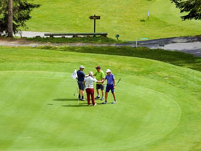 Four golfers on a green, talking and shaking hands under sunny skies, with lush grass all around.