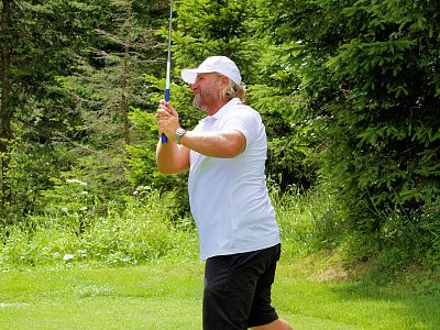 Man in white shirt and cap swings a golf club on a green lawn, with dense trees in the background. He's focused on the game amidst the serene nature.