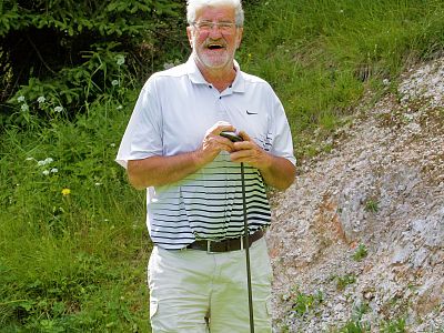 A man with a beard smiling and holding a golf club stands in a green grassy area. He wears a white and striped polo shirt and light-colored pants.