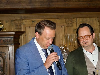 Zwei Männer stehen in einem eleganten Raum. Der Mann links spricht in ein Mikrofon, elegant gekleidet in einem blauen Anzug. Der andere trägt ein grünes Jackett.