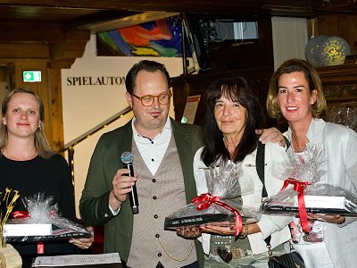 A group at an event, one person speaks into a microphone, others hold wrapped gifts. They stand together, smiling, in a warmly lit room with awards.