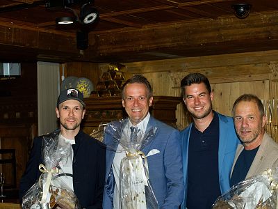 Four men in suits, two holding gift baskets, standing together in a wooden room with warm lighting.