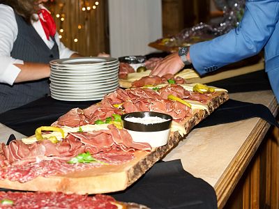 A charcuterie board with meats, garnishes, and a dip bowl on a wooden table. Several people are serving themselves at an indoor event.