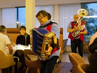 Two musicians in traditional outfits play accordion and brass instruments in a restaurant as diners watch and enjoy the music.