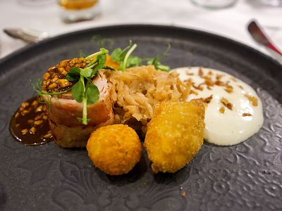 A gourmet dish with rolled meat, herbs, creamy sauce, crispy croquettes, and sauerkraut on a dark plate.