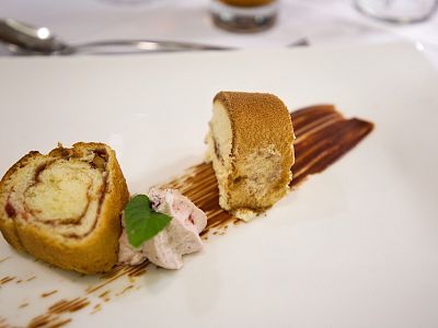 Two slices of rolled cake on a white plate with a streak of sauce, garnish leaf, and creamy dollop, elegantly presented with silverware in the background.