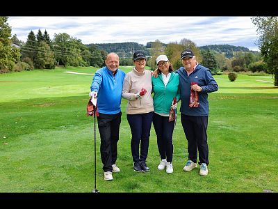 Four people smiling on a green golf course, holding clubs, wearing casual golf attire. Trees and hills are visible in the background under a cloudy sky.