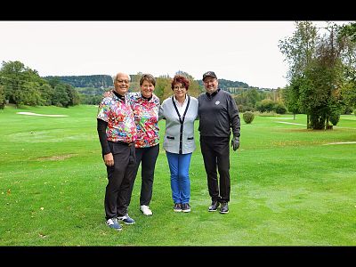 Four people stand on a green golf course with trees and hills in the background, wearing casual clothing. The weather appears overcast.