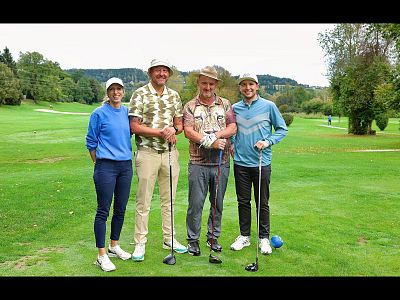 Four people stand on a golf course with trees in the background, holding golf clubs and wearing casual attire and hats, enjoying a bright, clear day.