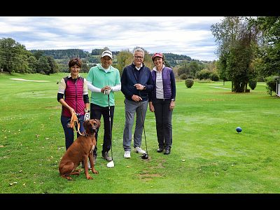 Four people and a dog stand on a sunny golf course, holding golf clubs. They're surrounded by trees and greenery, capturing a moment of leisure and camaraderie.