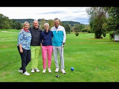 Vier Personen stehen lächelnd auf einem sonnigen Golfplatz. Sie tragen lässige, sportliche Kleidung und haben einen Golfschläger dabei.