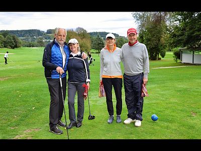 Four people posing on a golf course, wearing casual sports attire and holding golf clubs. The grassy field and trees are visible in the background.