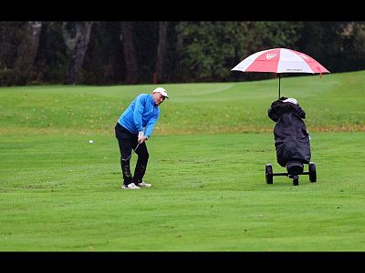 Golfer in blue jacket prepares to swing on green course; rain umbrella covers nearby golf cart.