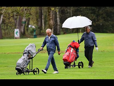 Two men on a golf course, one holding a white umbrella. Both push golf trolleys with bags on a rainy day, greenery and trees in the background.