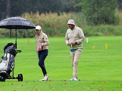 Two people on a wet golf course under an umbrella, dressed for rain. Golf bags are on the green. Trees and grass are in the background.