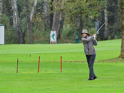 A golfer in a patterned jacket and hat swings a club on a green golf course with trees in the background. It's a cloudy day with lush greenery around.