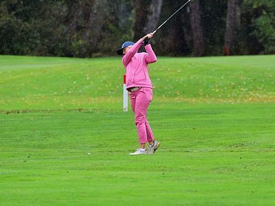 A child in pink clothing swings a golf club on a green field, surrounded by tall trees, on a cloudy day.