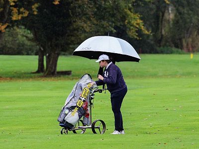 A person wearing a hat stands on a golf course under an umbrella, pushing a golf trolley. The background shows green grass and trees on a rainy day.